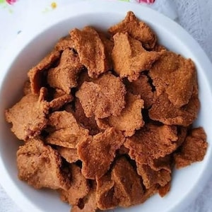 May include: A white bowl filled with a pile of brown, irregularly shaped, crispy snacks. The snacks have a textured surface and appear to be fried or baked. The bowl is set against a white background.