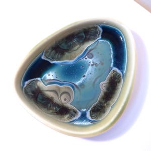 Puede incluir: Un plato de cerámica abstracto de forma libre. El interior presenta una mezcla de azules, verdes y tonos blanquecinos, creando un efecto marmolado. El borde es de un color crema suave. El plato mide aproximadamente 20 cm de largo.