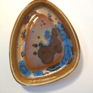 Puede incluir: Un plato triangular de cerámica con acabado brillante. El plato presenta un borde marrón y un centro con una mezcla de tonos marrones, naranjas y azules. El interior tiene un patrón abstracto único con formas circulares.