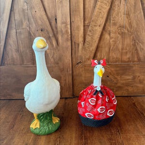 May include: Two decorative goose figurines. One is a white goose with a yellow beak and feet, standing on green grass. The other goose is wearing a red dress with white text and a red bow.