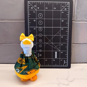 May include: A white ceramic goose figurine wearing a green and yellow patterned fabric dress and a yellow polka dot bow on its head, positioned next to a black measuring tool showing a height of approximately 4 inches for the goose.