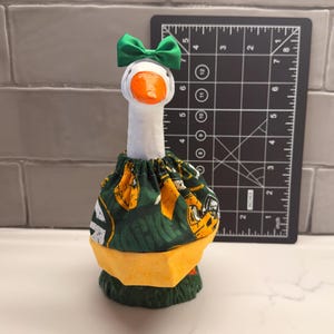 May include: A white goose figurine with an orange beak, wearing a green fabric bow on its head and a green and yellow patterned fabric outfit. A black measuring tool is positioned behind the goose, showing a height of approximately 6 inches.