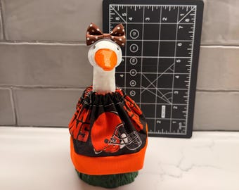 May include: A white ceramic goose figurine wearing a brown and white polka dot bow and a black fabric outfit featuring orange football helmets and the text BROWNS. A black ruler shows the figurine stands approximately 8 inches tall.