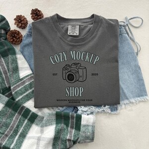 May include: A gray t-shirt with the text "COZY MOCKUP SHOP" and a camera graphic. The shirt is paired with denim shorts and a green and white plaid shirt. Decorative pinecones are scattered around the clothing.