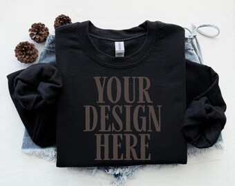 May include: A black crewneck sweatshirt with the text "YOUR DESIGN HERE" in a brown font. The sweatshirt is displayed on a pair of denim shorts, with three pinecones to the left. The sweatshirt is a Gildan brand.
