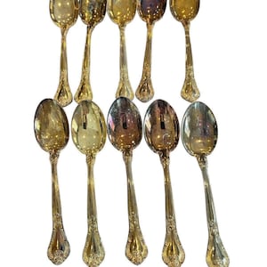 May include: A collection of ten antique gold-colored spoons. Each spoon features an oval bowl and a long, ornate handle with decorative detailing. The spoons are arranged on a white background, showcasing their reflective surfaces.