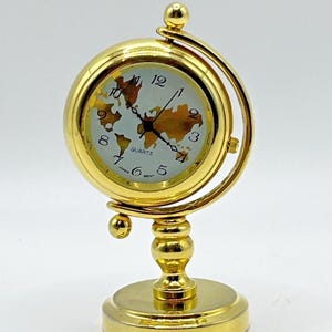 May include: A gold-toned metal desk clock featuring a spherical casing mounted on a pedestal base. The clock face displays a world map graphic in gold, black numerals, and thin black hands. The text QUARTZ and JAPAN MOVT are printed on the face.