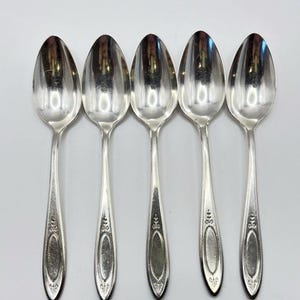 May include: Five silver spoons arranged in a row. Each spoon has an oval bowl and a long, slender handle with decorative detailing. The spoons are reflective, showing the surrounding environment.
