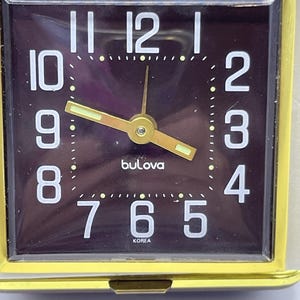 May include: A square Bulova travel alarm clock with a gold-tone metal frame and a dark purple face. The clock features white Arabic numerals, gold-tone hands with luminous inserts, and the text Bulova and KOREA printed on the face.