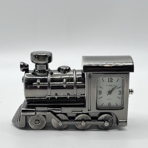 May include: A metallic gunmetal gray locomotive train clock with a rectangular white face featuring black numerals 3, 6, 9, and 12. The Timex quartz clock is integrated into the cab of the train, which has four wheels and a front cowcatcher.