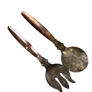 May include: A vintage salad serving set featuring a spoon and a fork. The spoon has a rounded bowl, and the fork has three tines. Both utensils have ornate silver-colored handles with copper-colored ends.