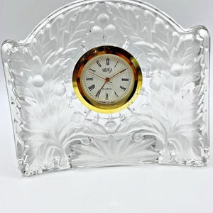 May include: A clear glass mantel clock with a frosted finish featuring a raised leaf and berry pattern. The clock face is white with black Roman numerals, gold-tone hands, and the text MIKASA QUARTZ printed in the center, surrounded by a gold rim.