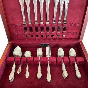 May include: A wooden silverware chest with a red velvet interior, showcasing a complete set of polished silverware. The set includes knives, forks, and spoons, all with ornate handle designs. The chest is open, revealing the neatly arranged utensils.