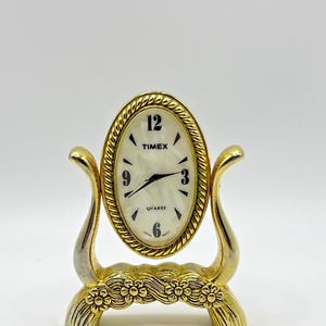May include: An oval-shaped gold-tone desk clock with a rope-textured border and a white pearlescent face. The clock features black numerals at 12, 3, 6, and 9, black hands, and the text TIMEX, QUARTZ, and JAPAN MOVT. It rests on a decorative floral base.