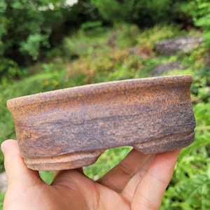 Może przedstawiać: Płytka, owalna doniczka ceramiczna do bonsai o teksturowanym wykończeniu w odcieniach ziemistego brązu i antracytowej szarości. Zewnętrzna powierzchnia ma szorstki, matowy wygląd z widocznymi poziomymi żłobieniami i grubym, lekko wywiniętym brzegiem.