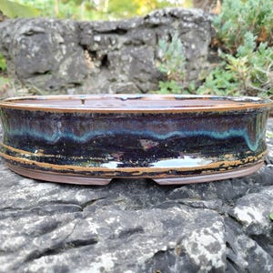 Peut inclure: Un pot à bonsaï rond et peu profond en céramique avec un glaçage bleu foncé et marron. Le pot a une forme large et peu profonde avec un bord légèrement évasé et une surface texturée. Le glaçage a une finition brillante avec des stries turquoise et marron.
