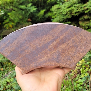 May include: A fan-shaped ceramic object with a textured, mottled brown and rust-colored surface. The piece features a curved top edge and a slightly concave bottom edge, displaying a weathered, earthy appearance with subtle variations in tone.