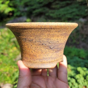 May include: A small, round, flared ceramic pot with a textured, speckled brown and tan finish. The exterior surface features subtle dark brown streaks and small raised bumps, with a wide, flat rim and a tapered base designed for planting.