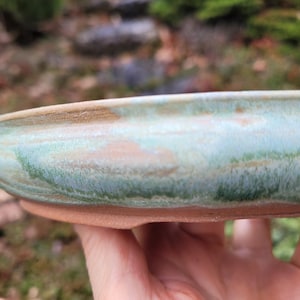 May include: A handmade ceramic bonsai pot with a rounded shape. The pot has a light green and brown glaze and a reddish-brown base. The pot is held in a hand, with a blurred outdoor background.
