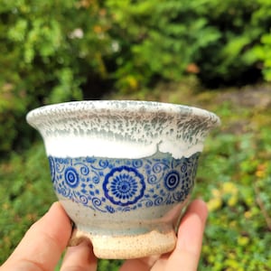 May include: A ceramic bowl featuring a flared rim with a white and gray speckled glaze. The midsection displays a blue floral pattern on a light gray base, while the base is unglazed, textured, and tan in color with small feet.