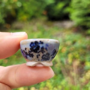 Può includere: Una ciotola in ceramica in miniatura con base azzurra, motivi floreali blu scuro e un piccolo piedistallo. La ciotola presenta un bordo liscio e arrotondato e una consistenza punteggiata su tutta la superficie, con un design delicato.