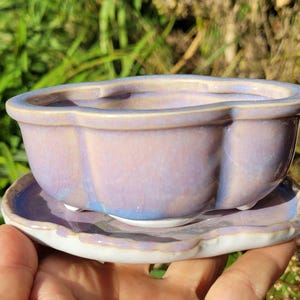 May include: A ceramic bonsai pot with a scalloped rim and a matching saucer. The pot features a glossy, iridescent lavender and light blue glaze with a smooth finish. It is supported by small, rounded feet and has a white base.