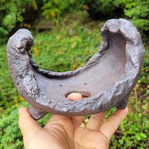 Peut inclure: Un pot à bonsaï en céramique gris foncé, façonné à la main, avec une texture rugueuse et irrégulière et un bord ondulé. Le récipient présente un large bassin ouvert, un petit trou de drainage au fond et trois pieds courts et robustes.