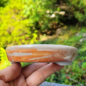 May include: A small, handmade ceramic dish with a rounded shape. The dish features a cream-colored top with orange horizontal stripes and a white base. The dish is held in a hand against a blurred green background.