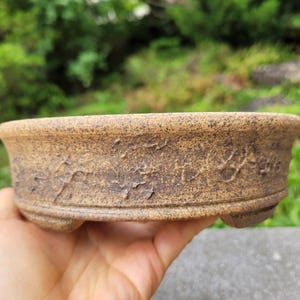 May include: A round, shallow ceramic bonsai pot with a textured, speckled tan and brown finish. The exterior features a rough, weathered surface with subtle carved markings along the side. The pot is supported by small, integrated feet at the base.