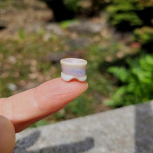 May include: A small, cylindrical ear plug with a flared design, featuring a smooth, iridescent finish that shifts between soft white, pale lavender, and subtle blue tones, with a concave centre and a rounded, notched base edge.