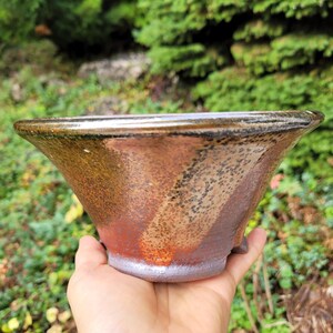 May include: A wide, flared ceramic bowl featuring a variegated glaze with earthy tones of brown, rust-orange, and charcoal grey. The exterior surface displays a speckled texture with a smooth, dark rim and a lighter, muted base.