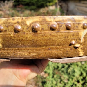 May include: A handmade, ceramic bowl with a textured, earthy appearance. The bowl is a warm, golden-brown color with a series of raised, rounded embellishments along the rim. The bowl's base is a lighter brown, and the overall design suggests a rustic, artisanal aesthetic.