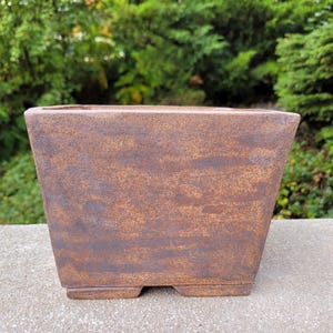 Peut inclure: Un pot de fleurs carré en céramique avec une finition texturée et tachetée de couleur marron et rouille foncé. Le pot présente une large ouverture, des côtés légèrement effilés et une base plate avec de petits pieds, offrant un aspect terreux.