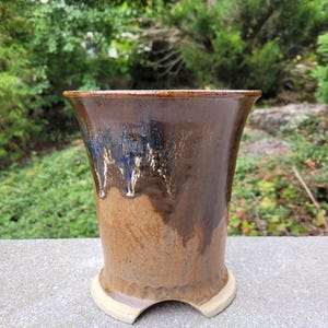 Op de afbeelding: Een handgemaakte keramische vaas met een uitlopende bovenkant en een uniek basisontwerp. De vaas heeft een mix van bruine, blauwe en crèmekleuren, met een glanzende afwerking. De buitenkant heeft een getextureerd uiterlijk, wat bijdraagt aan de ambachtelijke charme.