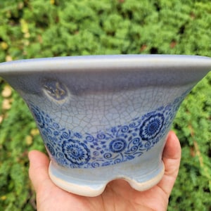 May include: A light blue ceramic bonsai pot with a flared rim and a wide base featuring a cutout. The exterior surface displays a fine crackle glaze finish and a decorative band of dark blue floral patterns encircling the middle section.