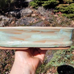 Könnte beinhalten: Ein rechteckiger Bonsai-Topf aus Keramik mit hellgrüner und brauner Glasur. Der Topf hat ein breites, flaches Design mit einer integrierten Untertasse. Die Kanten sind leicht nach außen gewölbt. Der Topf ist für Bonsai-Bäume konzipiert.