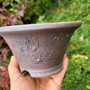 May include: A dark brown ceramic bonsai pot with a wide, flared rim and a textured, weathered surface. The exterior features a rough, uneven finish with raised, organic patterns and a sturdy base with small feet for elevation.