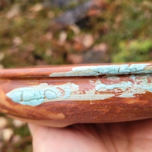 Könnte beinhalten: Eine handgefertigte, braune Keramikschale mit einem türkisfarbenen, strukturierten Design am Rand. Die Schale hat ein rustikales Aussehen mit sichtbaren Unvollkommenheiten und Farbvariationen. Die Schale wird wahrscheinlich zum Servieren oder Ausstellen verwendet.