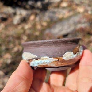 Könnte beinhalten: Ein kleiner, handgefertigter brauner Bonsai-Topf aus Keramik mit strukturierter Oberfläche. Der Rand ist dunkler braun, und die Basis hat hellblaue und weiße Akzente. Der Topf wird in einer Hand gehalten, mit verschwommenem Naturhintergrund.