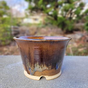 May include: A ceramic planter with a flared rim and a tapered body. The exterior features a glossy glaze in shades of deep brown and amber, with a textured, dripped effect near the base revealing a lighter, unglazed tan clay foot.