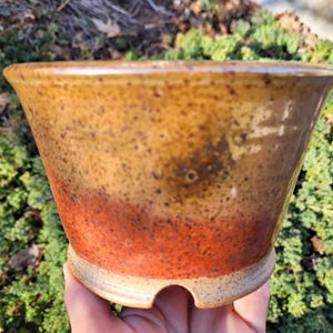 May include: A handmade ceramic planter with a speckled, gradient design. The top is a warm, golden-brown, transitioning to a reddish-orange at the base. The planter has a flared shape and a light-colored, textured base.