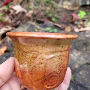 May include: A handcrafted ceramic cup with a flared rim and a textured base. The cup displays a colour gradient, shifting from a light tan at the base to a warm orange-brown at the top. Small dark speckles dot the surface.