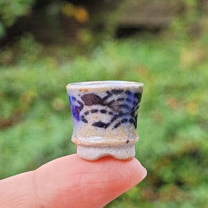 Peut inclure: Un pot en céramique miniature avec une base beige et une finition brillante, orné de motifs abstraits peints à la main en bleu foncé et noir sur l'extérieur. Le petit récipient présente un bord évasé et une base à pied avec une encoche.