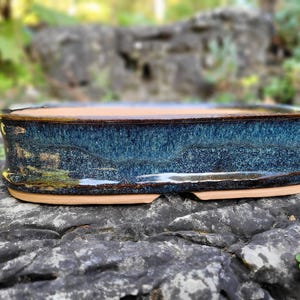 Könnte beinhalten: Handgefertigter, ovaler Bonsai-Topf. Der Keramiktopf hat eine strukturierte, dunkelblaue Glasur mit braunen und beigen Akzenten. Der Topf steht auf einem grauen Felsen.