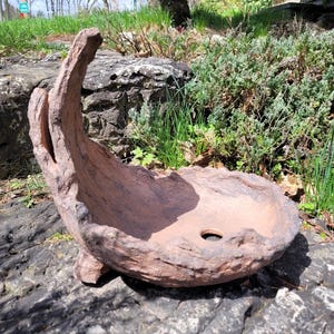 May include: A brown, textured, decorative planter with an unusual, organic form. The planter features a curved, upward-reaching section and a rounded bowl with a drainage hole. The planter is made from a material that resembles wood.