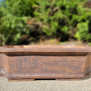 May include: A rectangular, brown bonsai pot with a textured surface. The pot has a wide, shallow design with a slightly flared rim and decorative corners. The pot is made of a durable material.