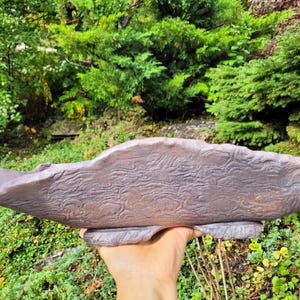 May include: A long, shallow, hand-formed ceramic tray with a dark, matte, purplish-brown finish. The surface features a distinct, raised, swirling, wood-grain-like texture across its entire length, with an irregular, organic, wavy rim edge.