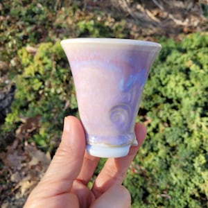 Peut inclure: Une tasse en céramique faite à la main avec un bord évasé et un petit pied. La tasse présente un mélange de teintes lavande, rose et bleu, avec un motif tourbillonnant subtil. L'extérieur a une finition brillante. La tasse est contre un fond de feuillage vert.