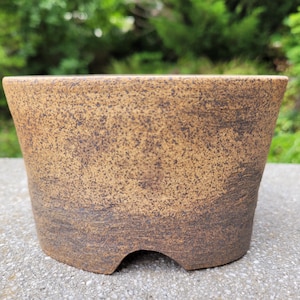 May include: A round ceramic bonsai pot with a tapered silhouette and a wide opening. The exterior features a textured, speckled tan finish that transitions into a darker, earthy brown tone toward the base, which includes a small arched cutout.