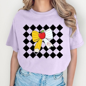 May include: A light purple t-shirt featuring a school-themed graphic. The graphic includes a yellow pencil, a red apple, and a white and blue bow against a black and white checkered background. The text "Proverbs 31:26" is visible.